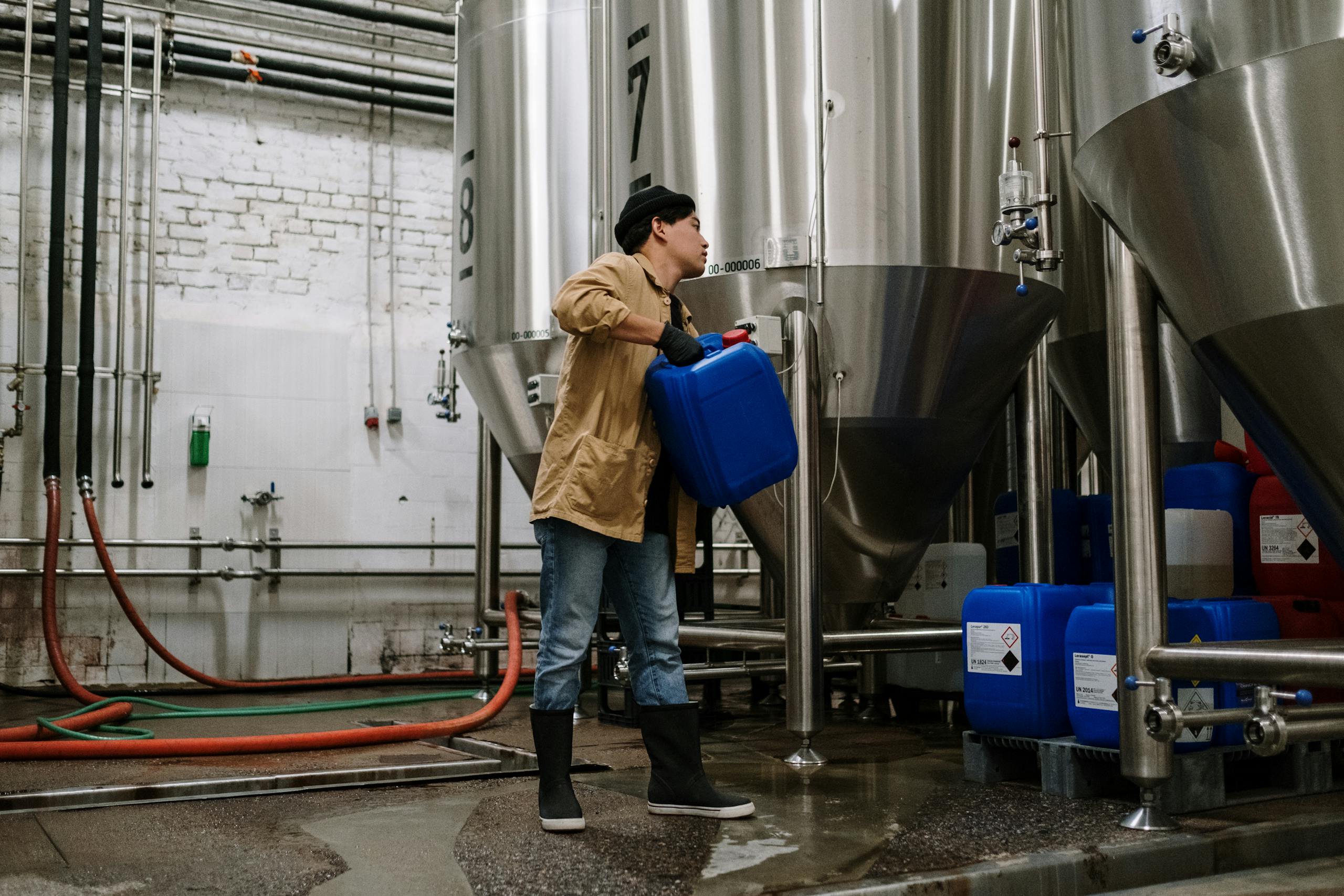 Man handling blue container in a brewery with stainless steel tanks.