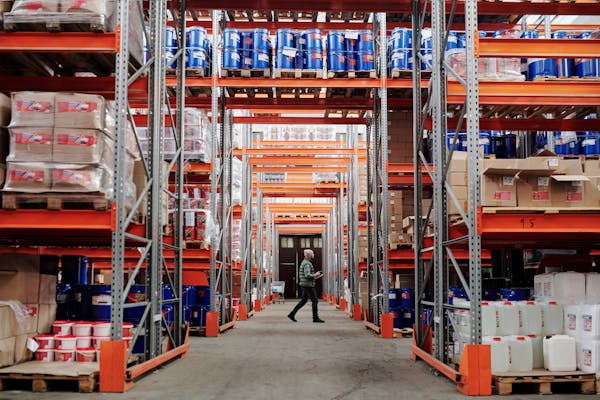 A man walking through a large industrial warehouse with stacked shelves filled with goods and products.