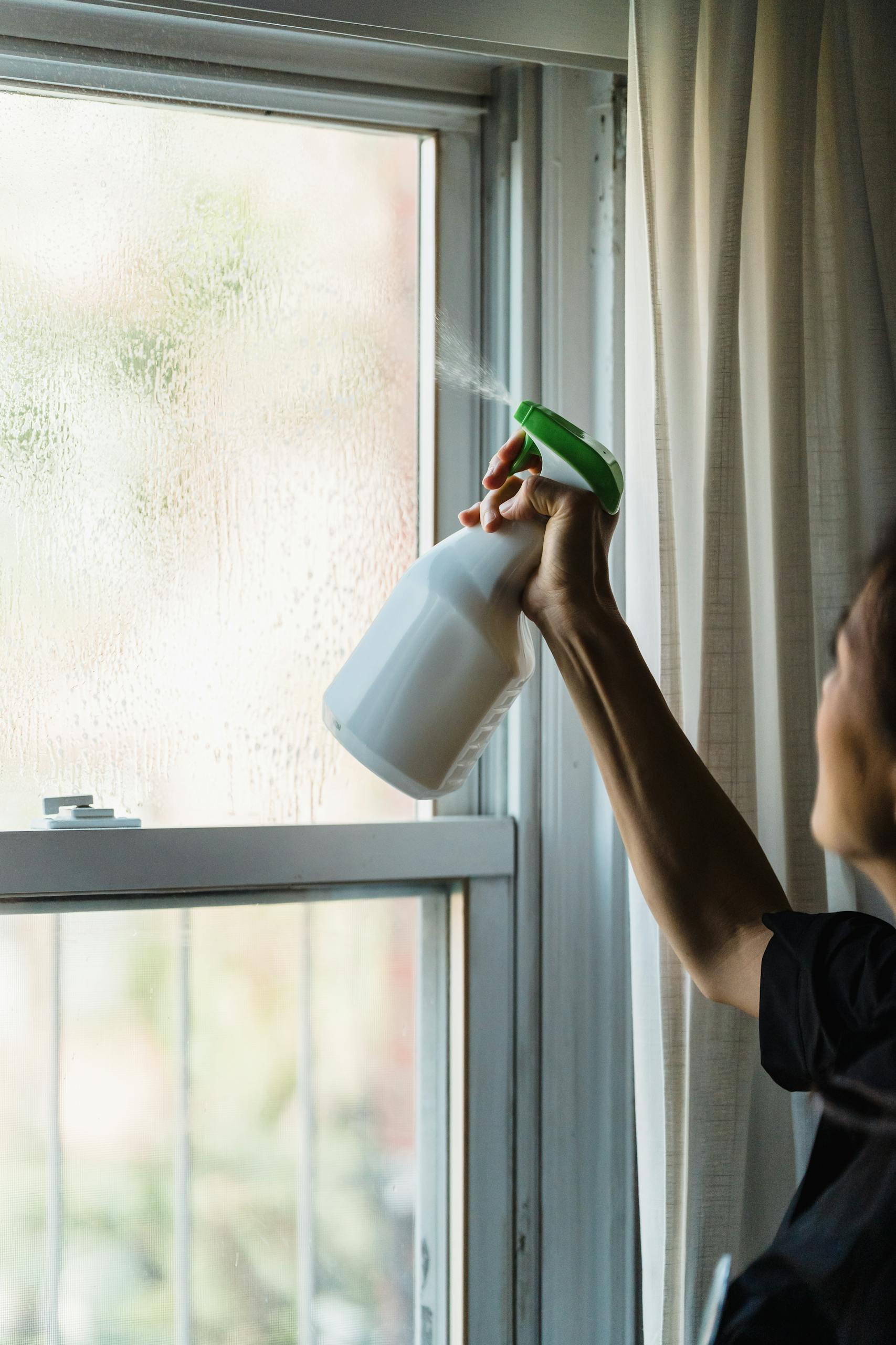 A person cleaning a window with a spray bottle indoors, emphasizing cleanliness and hygiene.
