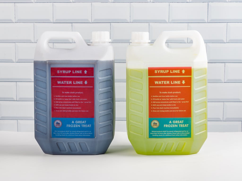 Two syrup containers for slush products against white tile background.