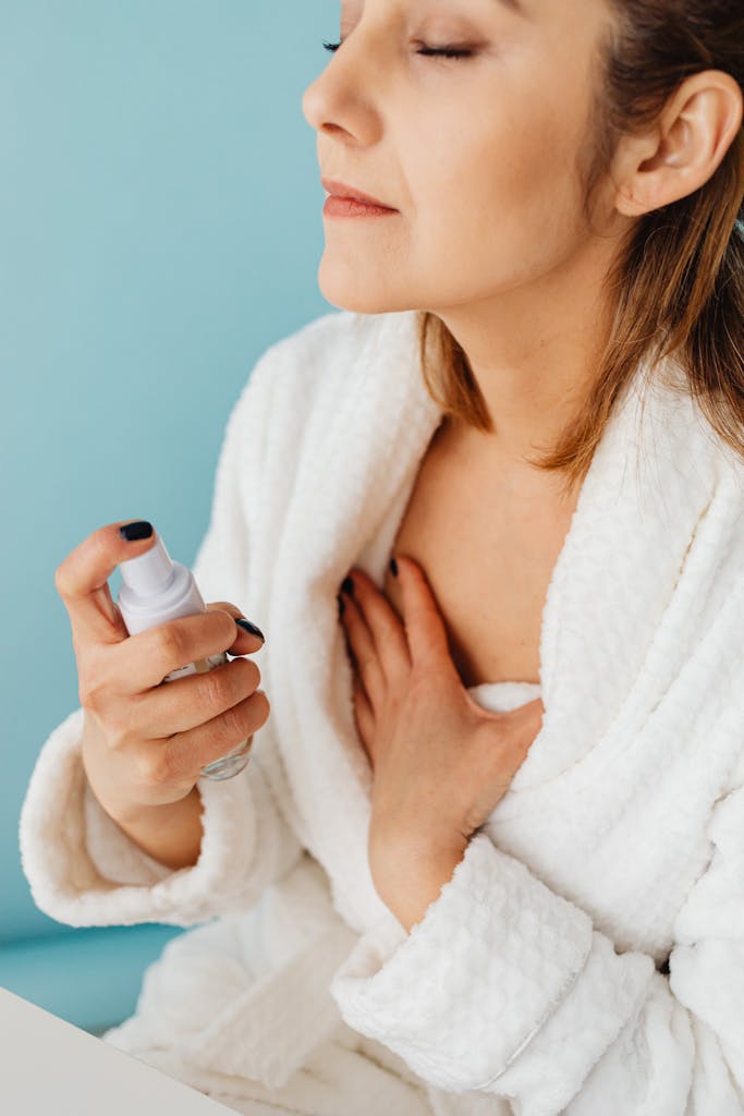 Woman in bathrobe applying skincare spray with eyes closed against blue background.